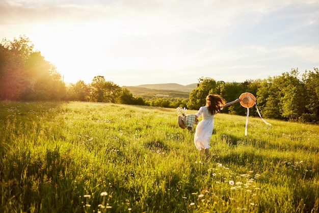 une femme en robe légère avec un chapeau dans les mains court loin dans le champ au coucher du soleil