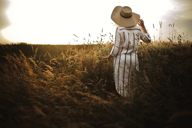 Photo femme en robe de lin et chapeau marchant dans les fleurs sauvages et les herbes au coucher du soleil lumière dorée dans la prairie d'été élégante fille rustique appréciant la soirée dans la campagne moment atmosphérique espace de copie