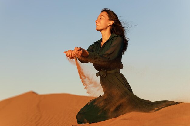 Femme en robe longue verte se dresse dans les dunes de sable du désert fond coucher de soleil orange jaune