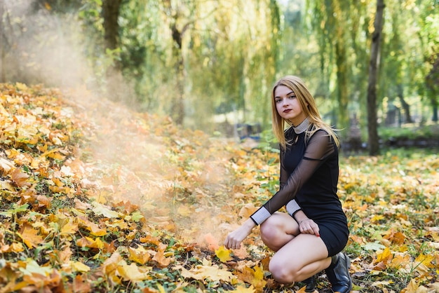 Femme en robe noire avec une bombe fumigène dans un parc d'automne posant sur des feuilles jaunes