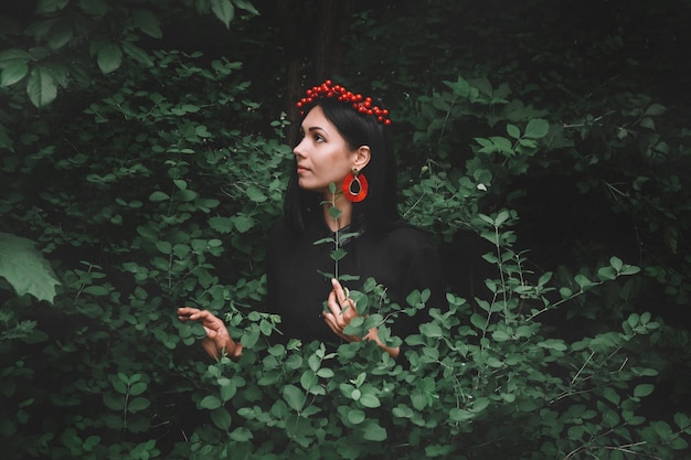 femme en robe noire et décorations rouges qui tient une brindille à la main sur fond de forêt