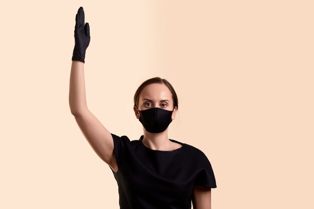 Femme en robe noire, masque facial et gants en latex a levé la main comme demander le droit de vote et sur un mur beige