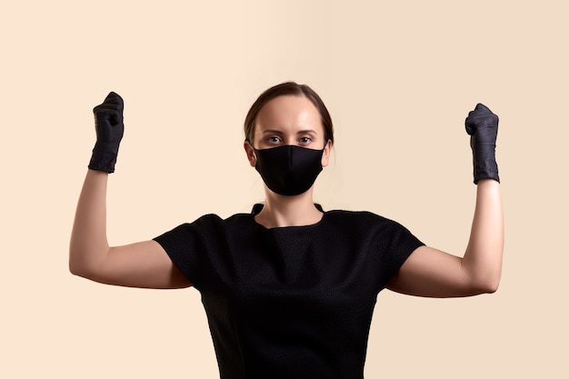 Femme en robe noire masque et gants a levé les mains avec les poings et sur le mur beige