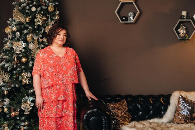 Une femme en robe rouge debout à côté d'un arbre de Noël dans un intérieur décoré pour célébrer Noël et le Nouvel An