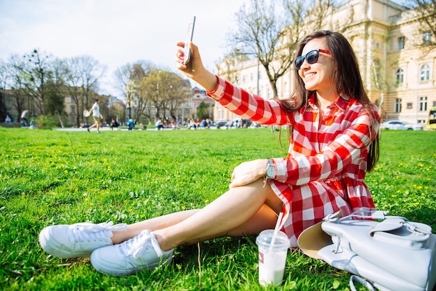 Femme en robe rouge s'asseoir dans le parc sur l'herbe verte avec smartphone