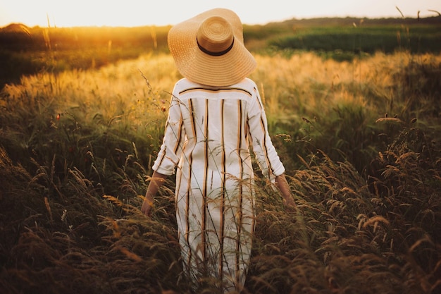 Femme en robe rustique et chapeau marchant dans les fleurs sauvages et les herbes au coucher du soleil lumière dorée dans la prairie d'été Moment authentique atmosphérique Fille élégante appréciant la soirée à la campagne