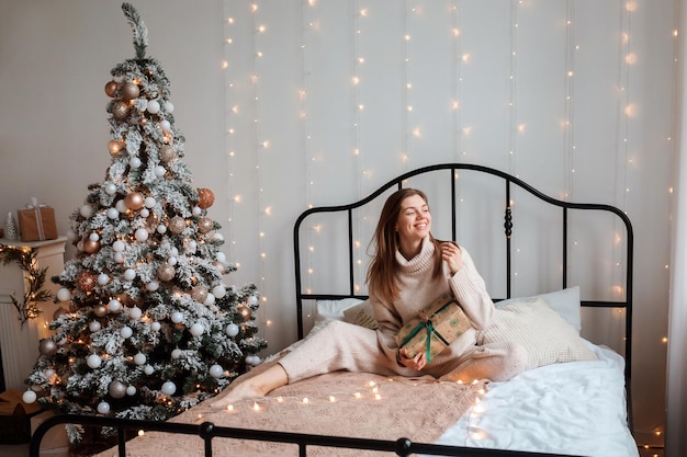 Une femme romantique est assise sur le lit avec un cadeau de Noël dans ses mains, à côté de l'arbre de Noël. Fille passant des vacances d'hiver à la maison.