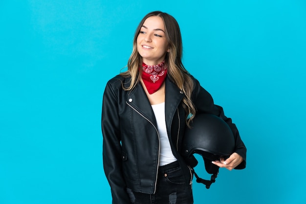 Femme roumaine tenant un casque de moto isolé sur fond bleu à la recherche sur le côté et souriant