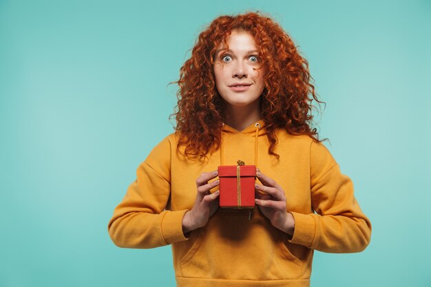 femme rousse affable des années 20 souriant et tenant une boîte présente isolée sur un mur bleu