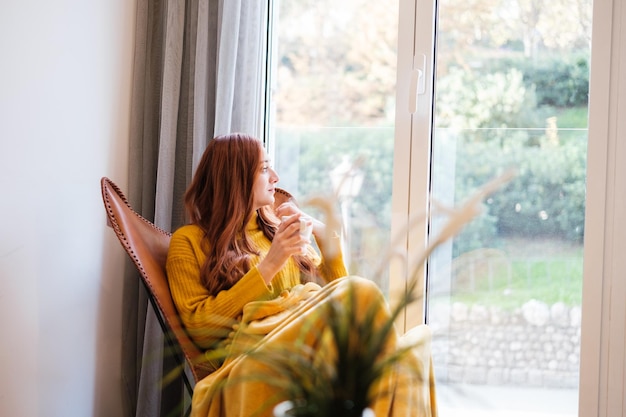 Femme rousse assise sur la chaise près de la fenêtre avec une tasse de café à la tranquillité réfléchie Concept détente repos