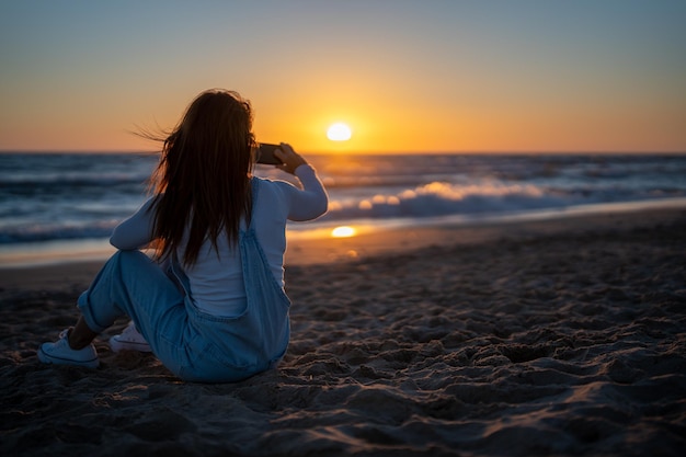 Femme rousse assise sur le sable d'une plage filmant une vidéo d'un coucher de soleil