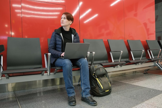 Une femme rousse aux cheveux courts en vêtements d'extérieur travaille sur un ordinateur portable à l'aéroport en le mettant sur ses genoux