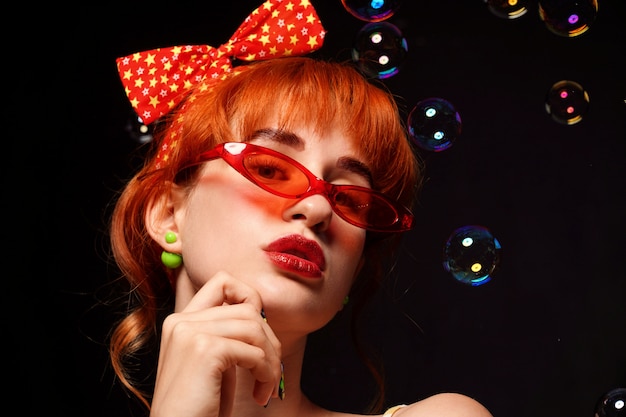 Femme rousse avec un bandeau lumineux et des lunettes rouges sur fond noir.