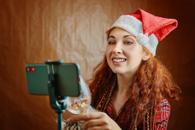 Femme rousse avec bonnet de Noel et dreadlocks clins de verre sur smartphone. Conversation virtuelle avec les parents et utilisation du chat vidéo. Appel vidéo de Noël.