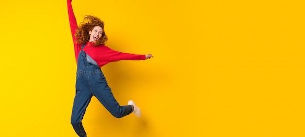 Femme rousse avec une combinaison sautant par-dessus un mur jaune isolé