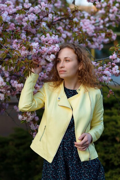 Une femme rousse dans une veste jaune regarde de côté debout près d'un magnolia en fleurs