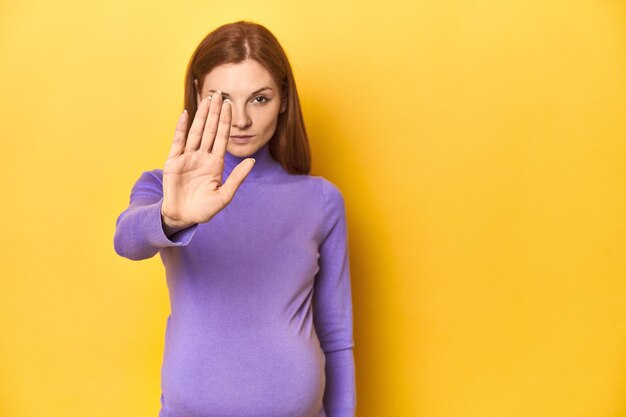 Femme rousse enceinte anticipant la maternité debout avec la main tendue montrant un panneau d'arrêt