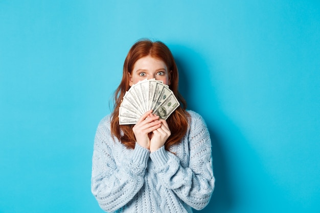 Femme rousse excitée couvrant le visage avec de l'argent, tenant des dollars et regardant la caméra heureuse, debout sur fond bleu.