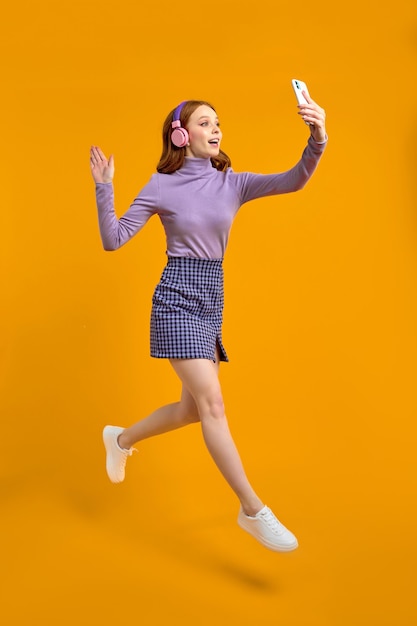 Une femme rousse excitée dans des écouteurs sautant en marchant et parlant au téléphone en ligne via une caméra dit bonjour ...