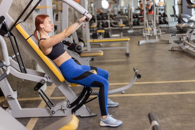 Une femme rousse en forme musclée entraîne les muscles de la poitrine dans une machine d'exercice à la salle de sport