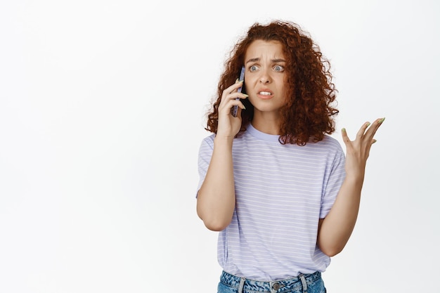 Photo une femme rousse frustrée reçoit un appel téléphonique bouleversant, parle sur un smartphone avec un visage inquiet et inquiet, debout sur fond blanc, reçoit de mauvaises nouvelles.