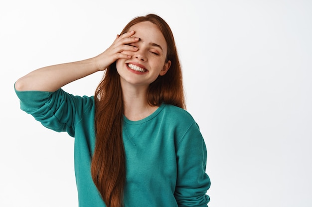 Femme rousse insouciante riant, souriante aux dents blanches, ferme les yeux et touche doucement le visage, se sentant libre et heureuse, debout sur blanc