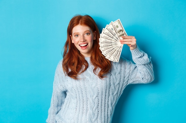 Femme rousse joyeuse en pull montrant des dollars, souriant heureux et tenant de l'argent, debout sur fond bleu.