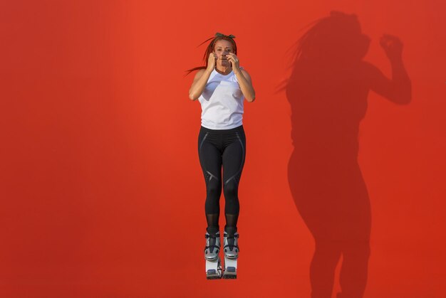 Femme rousse latine en vêtements de sport sautant avec les bras et les jambes fermés portant des bottes Kangoo Jumps sur fond orange. Concept de vie saine, d'activité et de sport