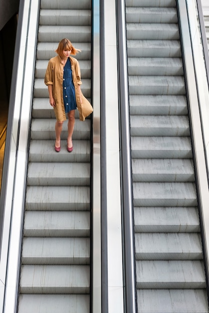 Femme rousse à la mode à cheval seule sur un long escalator descendant raide dans un immeuble de la ville ou un centre commercial dans une vue à distance avec fond
