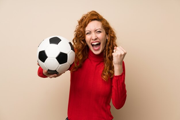 Femme rousse avec un pull à col roulé tenant un ballon de foot