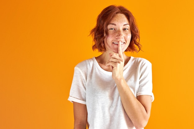 femme rousse a un secret, montrer garder le geste de silence, sourire en regardant la caméra sur orange