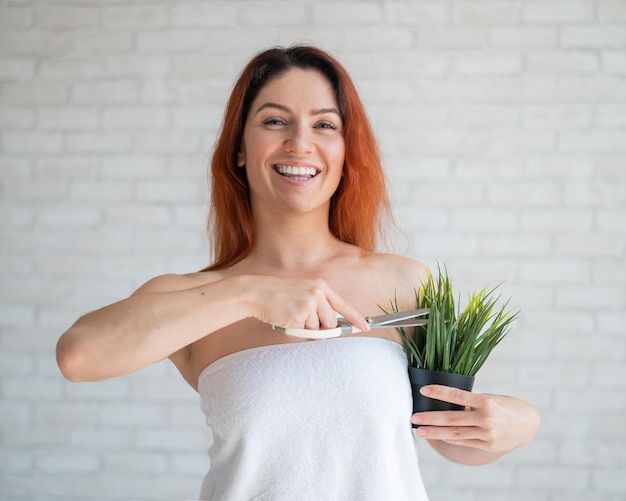 Une femme rousse souriante dans une serviette éponge blanche coupe une plante verte dans un pot avec des ciseaux Imitation de l'épilation des aisselles Concept d'épilation Procédure d'épilation de la végétation indésirable