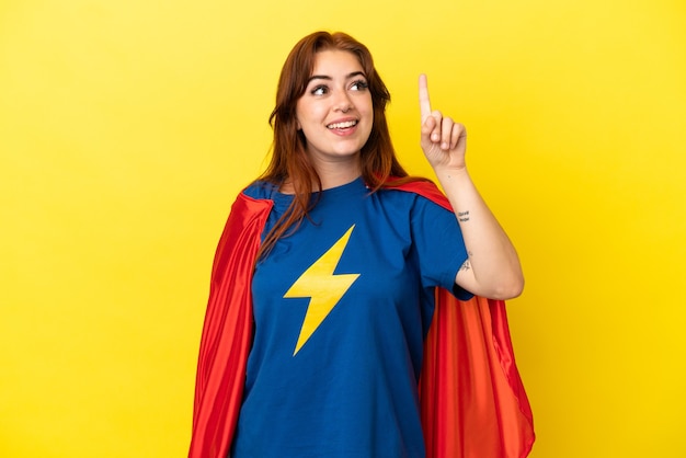 Femme rousse super héros isolée sur fond jaune dans l'intention de réaliser la solution tout en levant un doigt vers le haut