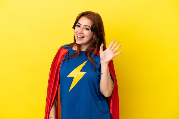 Femme rousse super héros isolée sur fond jaune saluant avec la main avec une expression heureuse