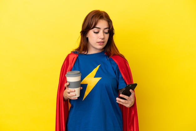 Femme rousse super héros isolée sur fond jaune tenant du café à emporter et un mobile