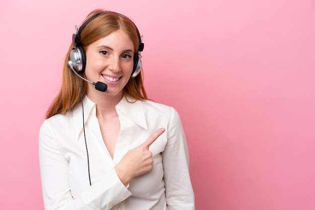 Femme rousse de télévendeur travaillant avec un casque isolé sur fond rose pointant vers le côté pour présenter un produit