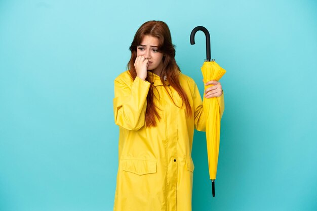 Femme rousse tenant un parapluie isolé sur fond bleu ayant des doutes