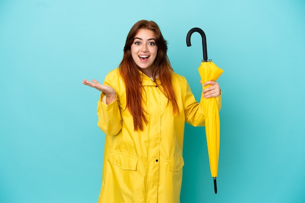 Femme rousse tenant un parapluie isolé sur fond bleu avec une expression faciale choquée