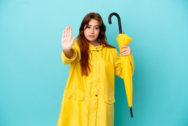 Femme rousse tenant un parapluie isolé sur fond bleu faisant un geste d'arrêt