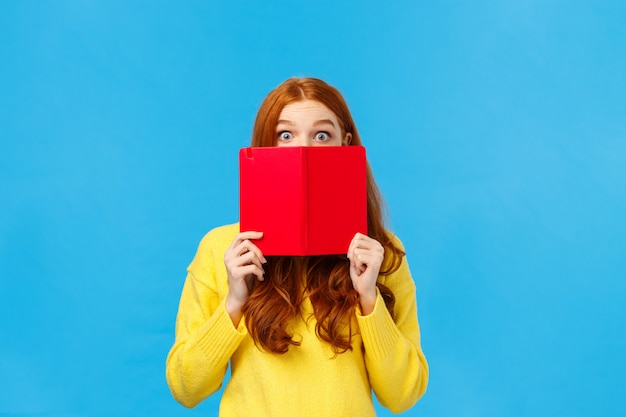 Femme rousse timide et mignonne comme lire des livres et écrire dans un journal, se cachant le visage derrière un cahier cool rouge