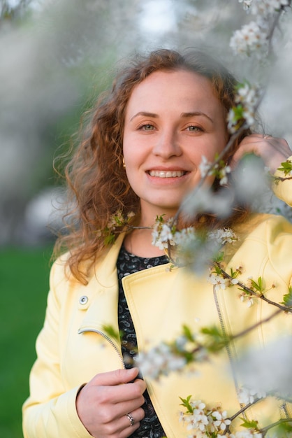 Une femme rousse en veste jaune regarde la caméra dans un jardin de printemps en fleurs