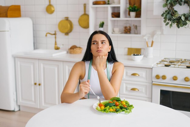 Femme avec un ruban à mesurer autour du cou est assise à une table avec un visage insatisfait devant une assiette de légumes Le concept d'une alimentation saine