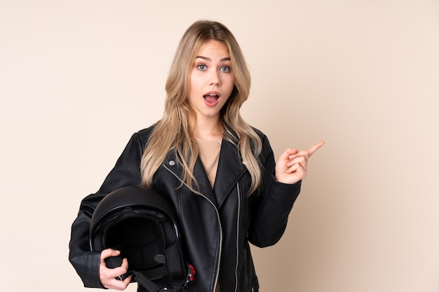 Femme russe avec un casque de moto isolé sur mur beige surpris et pointant le côté