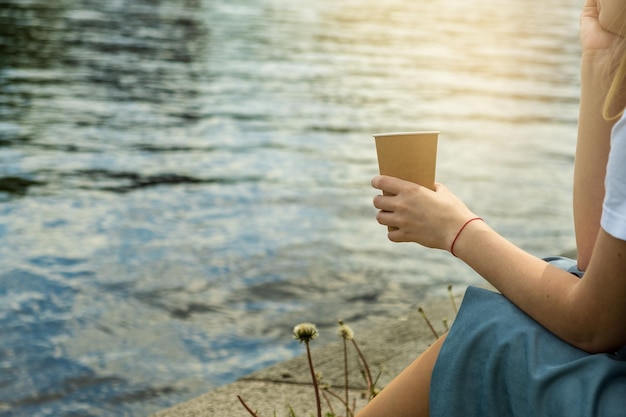 La femme s'assied sur le rivage de l'étang et tient la tasse ecofriendly