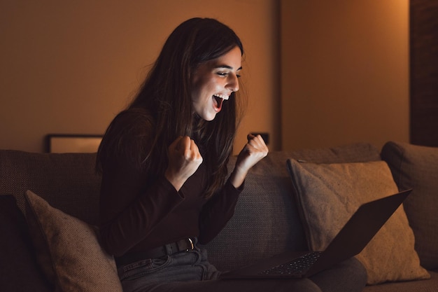 La femme s'assoit sur le canapé lit le courrier électronique sur l'ordinateur portable fait le oui le geste se sent heureux l'entrepreneur féminin obtient de grandes nouvelles d'affaires célèbre la croissance de la carrière avance la réalisation gagne le moment de la victoire de l'enchère la nuit xA