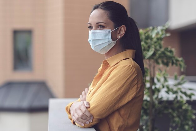 Photo femme s'auto-isolant à la maison pendant la pandémie de coronavirus, elle s'appuie sur le balcon et porte un masque facial