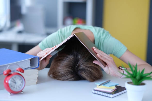 Une femme s'est couverte la tête avec un ordinateur portable et l'a posé sur une table sur le lieu de travail. Traitement du concept d'heures de travail