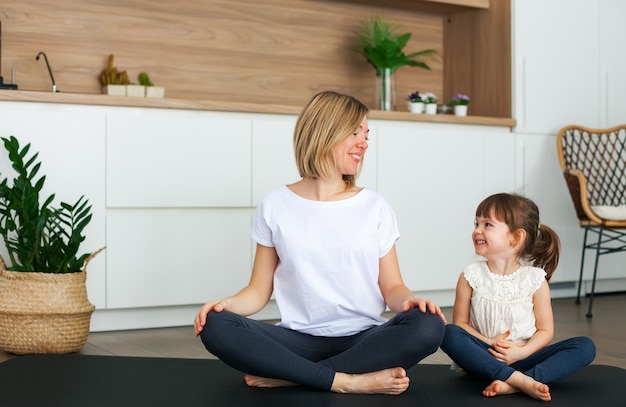 Femme et sa jolie petite fille sont assis dans une position du lotus tout en faisant du yoga ensemble à la maison