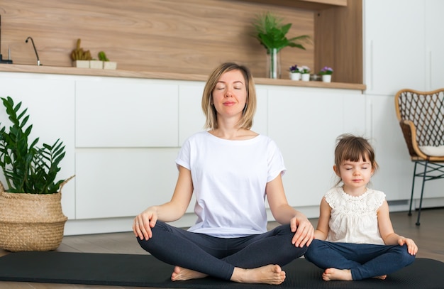 Femme et sa petite fille sont assis dans une position du lotus avec les yeux fermés tout en faisant du yoga ensemble à la maison