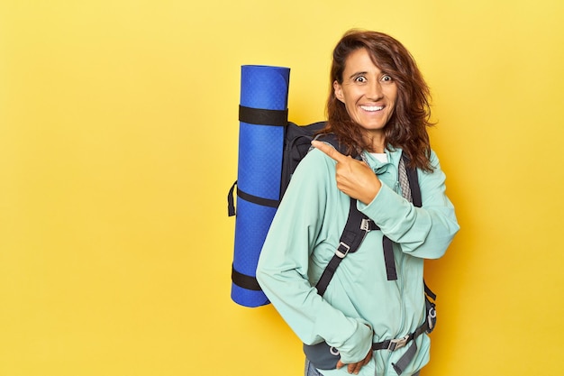 Femme avec sac à dos de randonnée et tapis sur jaune souriant et pointant de côté montrant quelque chose